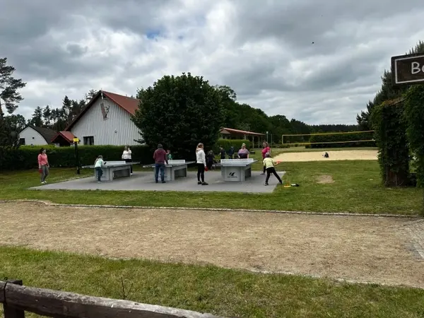 Mehrere Menschen spielen Tischtennis im Freien neben einem Volleyballfeld und Gebäuden.