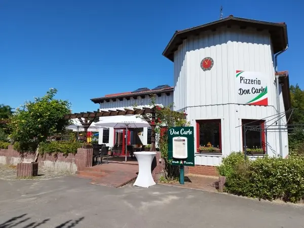 Außenansicht der Pizzeria Don Carlo mit Terrasse, Sonnenschirm und grüner Bepflanzung.